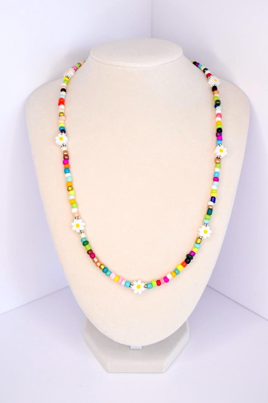 A colorful beaded necklace with white daisy flower accents. The necklace features a variety of small, square beads in shades of red, pink, yellow, green, blue, and black. The daisy flowers are evenly spaced throughout the necklace, adding a whimsical touch to the jewelry.