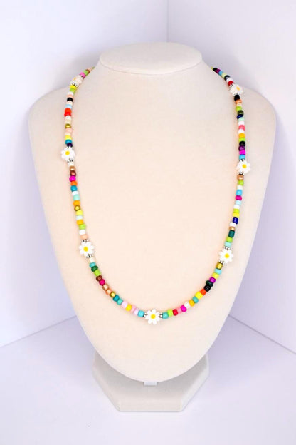 A colorful beaded necklace with white daisy flower accents. The necklace features a variety of small, square beads in shades of red, pink, yellow, green, blue, and black. The daisy flowers are evenly spaced throughout the necklace, adding a whimsical touch to the jewelry.