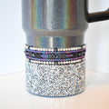 A midnight glitz Stanley tumbler with a black handle. The bottom section is covered in small, sparkling silver rhinestones. Above this is a band of beads in silver, purple, and black, with a central band of black arrow-shaped beads. The upper portion of the tumbler has a holographic, iridescent finish.