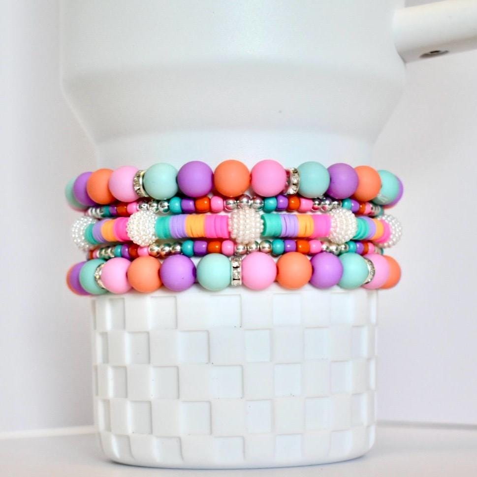 Stack of colorful beaded bracelets in shades of pink, orange, purple, and teal. The bracelets feature round beads and flat disc beads, with silver accents and white pearl-like embellishments. The bracelets are stacked on a white, textured container.