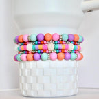 Stack of colorful beaded bracelets in shades of pink, orange, purple, and teal. The bracelets feature round beads and flat disc beads, with silver accents and white pearl-like embellishments. The bracelets are stacked on a white, textured container.