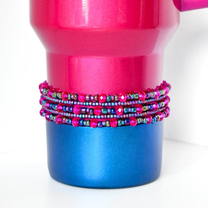 A pink and blue tumbler with a beaded wrap. The tumbler is primarily pink with a blue base. The beaded wrap is multi-colored, featuring pink, purple, and green beads. The beads are arranged in multiple rows around the tumbler, adding a decorative element.