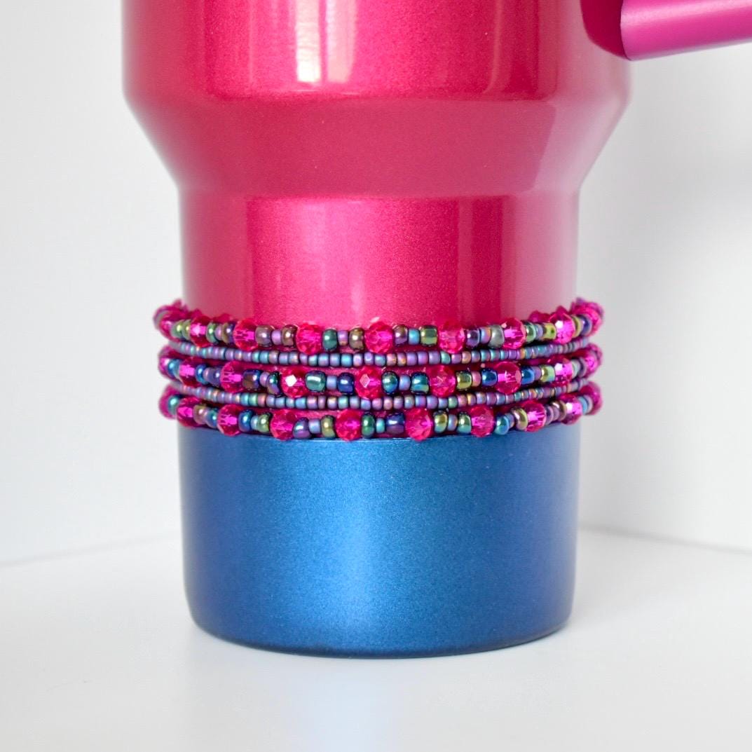 A pink and blue Berry truffle Stanley tumbler with a beaded wrap. The tumbler has a shiny pink top and a blue base. The wrap is made of multiple strands of small, colorful beads in shades of pink, purple, and green, adding a decorative element.