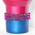 A berry truffle Stanley with a stack of matching bracelets. The top portion is a vibrant pink, while the base is a deep blue. Wrapped around the middle are multiple strands of beaded jewelry in shades of pink, purple, and iridescent green, creating a decorative accent.