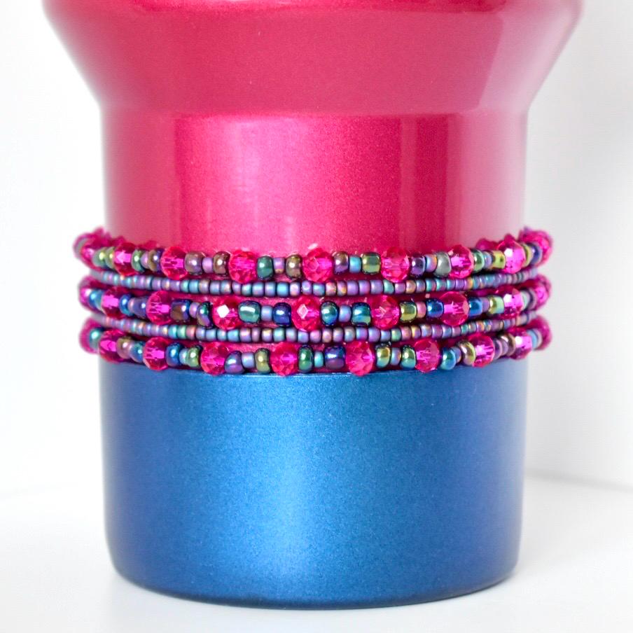 A berry truffle Stanley with a stack of matching bracelets. The top portion is a vibrant pink, while the base is a deep blue. Wrapped around the middle are multiple strands of beaded jewelry in shades of pink, purple, and iridescent green, creating a decorative accent.