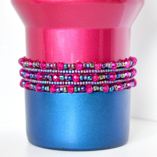 A berry truffle Stanley with a stack of matching bracelets. The top portion is a vibrant pink, while the base is a deep blue. Wrapped around the middle are multiple strands of beaded jewelry in shades of pink, purple, and iridescent green, creating a decorative accent.