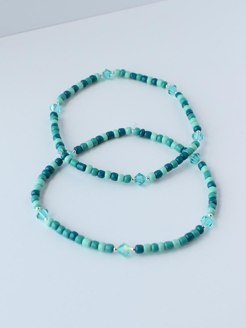 Two beaded bracelets in shades of teal and turquoise. The bracelets are made of small, round beads interspersed with faceted crystal beads. The bracelets are displayed on a white surface.