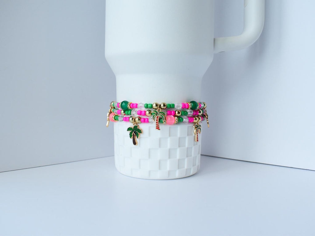 Colorful stackable bracelets featuring bright pink, green, and gold beads, rhinestone spacers, and playful palm charms, draped over a white mug for a chic display.