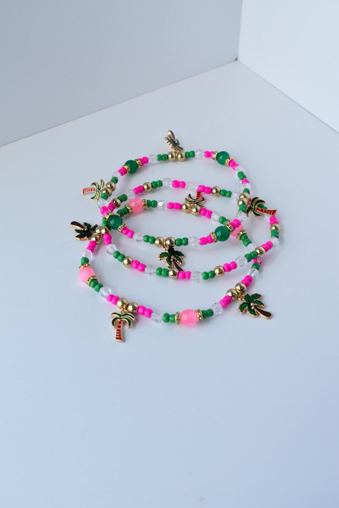 Tropical-themed bracelet set displayed loosely coiled; includes neon pink and emerald green beads, gold accents, clear glass beads, and playful palm tree charms for a beachy look