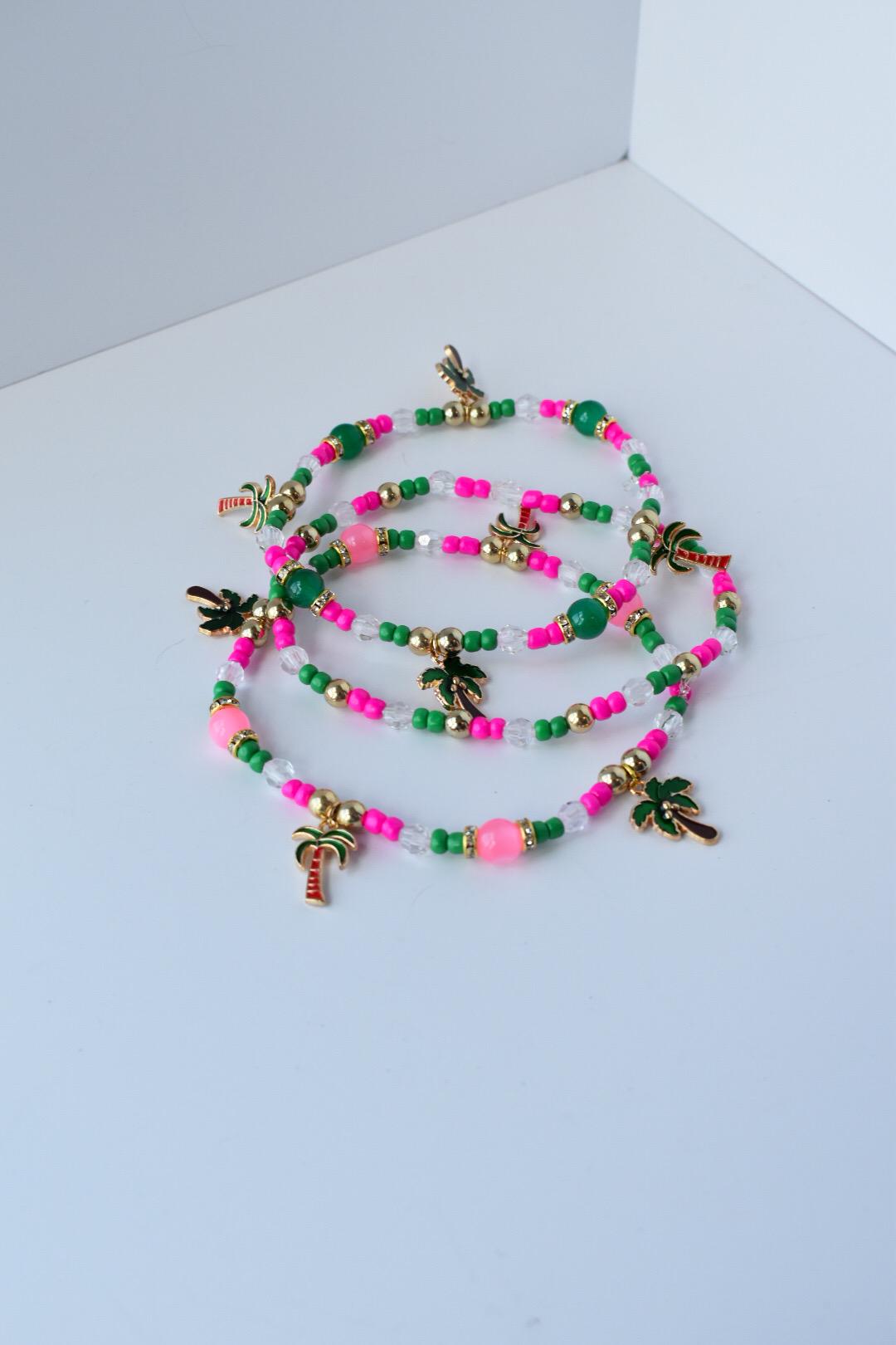 Tropical-themed bracelet set displayed loosely coiled; includes neon pink and emerald green beads, gold accents, clear glass beads, and playful palm tree charms for a beachy look