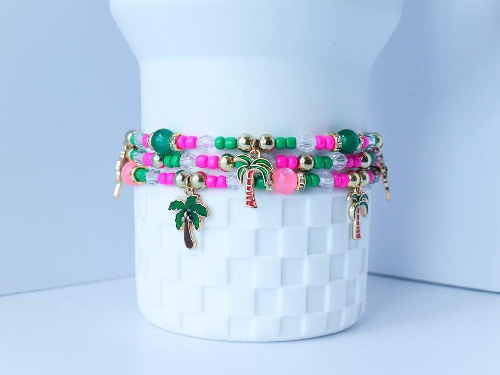 Handmade tropical beaded bracelets stacked with pink, green, and gold beads and dangling palm tree charms, shown wrapped around a white tumbler.
