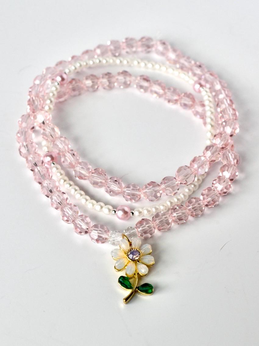 A pink beaded necklace with three strands. The top and bottom strands are made of faceted pink beads, while the middle strand features small white beads. A flower pendant with white petals, a gold center, and a purple gem hangs from the necklace.