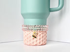 A light blue tumbler with a handle sits atop a pink, geometric patterned base. Three beaded bracelets in shades of pink and white are stacked around the tumbler's base. A charm with a white flower and green stem hangs from the bracelets.