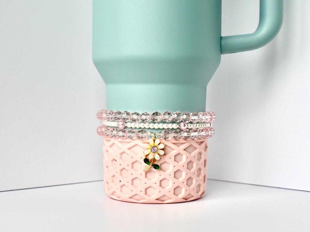 A light blue tumbler with a handle sits atop a pink, geometric patterned base. Three beaded bracelets in shades of pink and white are stacked around the tumbler's base. A charm with a white flower and green stem hangs from the bracelets.