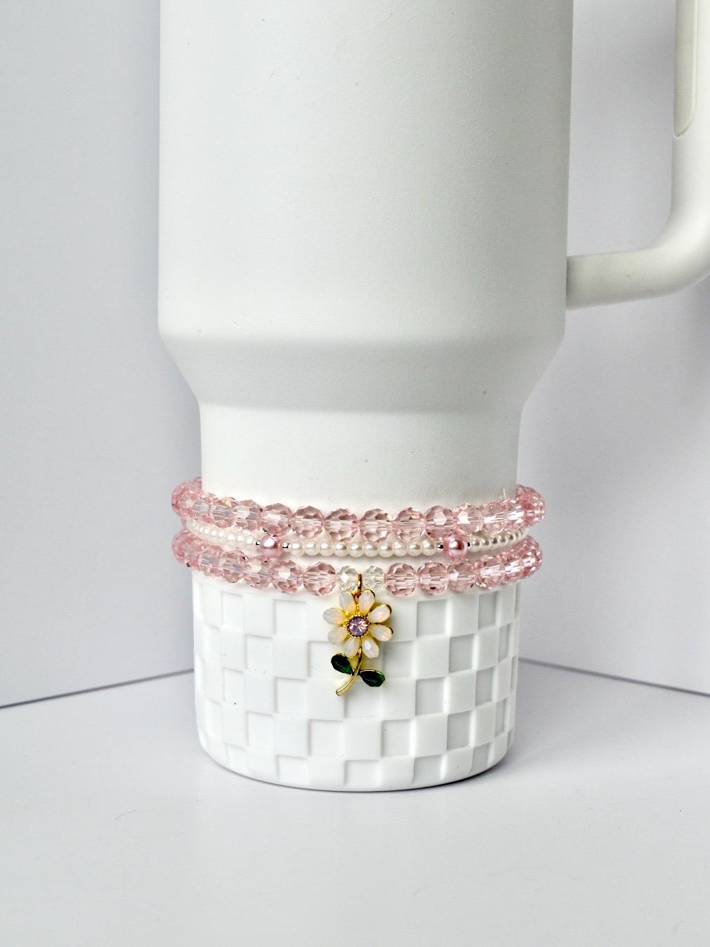 A white tumbler with a decorative pink beaded bracelet set around the base. The bracelets are made of pink beads and white pearls. A gold-colored charm with a pink and white flower with green leaves hangs from the bracelets.