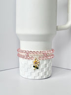 A white tumbler with a decorative pink beaded bracelet set around the base. The bracelets are made of pink beads and white pearls. A gold-colored charm with a pink and white flower with green leaves hangs from the bracelets.