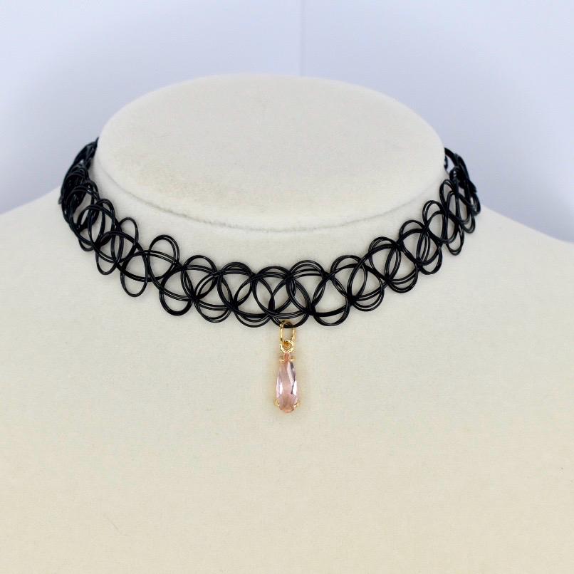 A black choker necklace with a repeating circular pattern. A teardrop-shaped, light pink gemstone pendant hangs from the center. The necklace is displayed on a white surface.