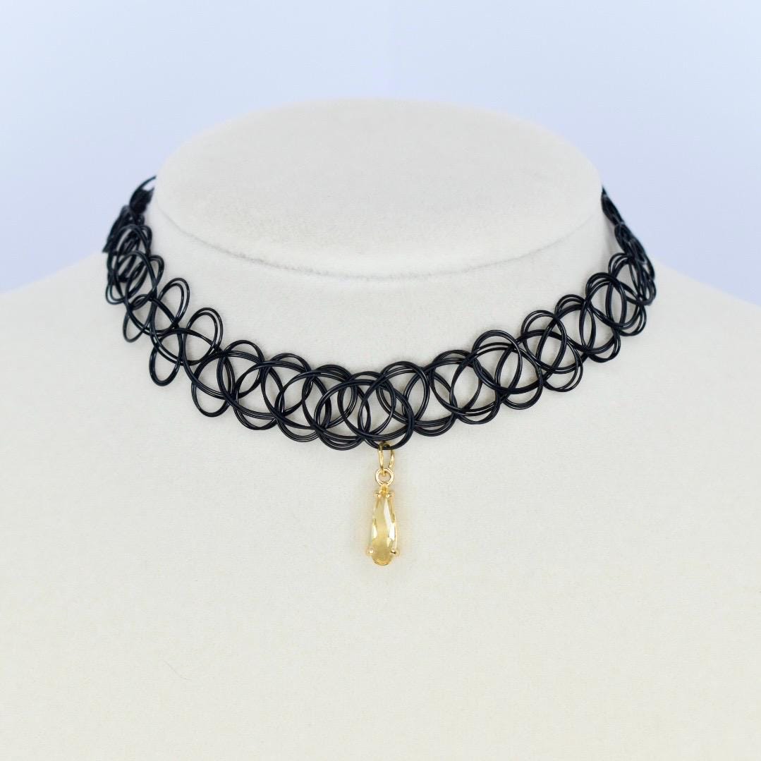 A black choker necklace with a pendant. The necklace is made of a series of interconnected circular loops. A gold-colored teardrop-shaped pendant hangs from the center of the necklace. The necklace is displayed on a white mannequin.