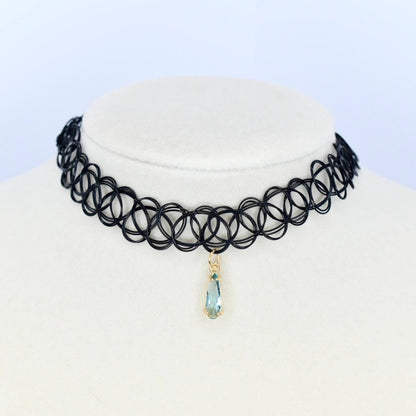 A black choker necklace with a pendant. The necklace is made of small, interconnected circles. A teardrop-shaped pendant hangs from the center, featuring a light blue gemstone set in a gold-colored metal.