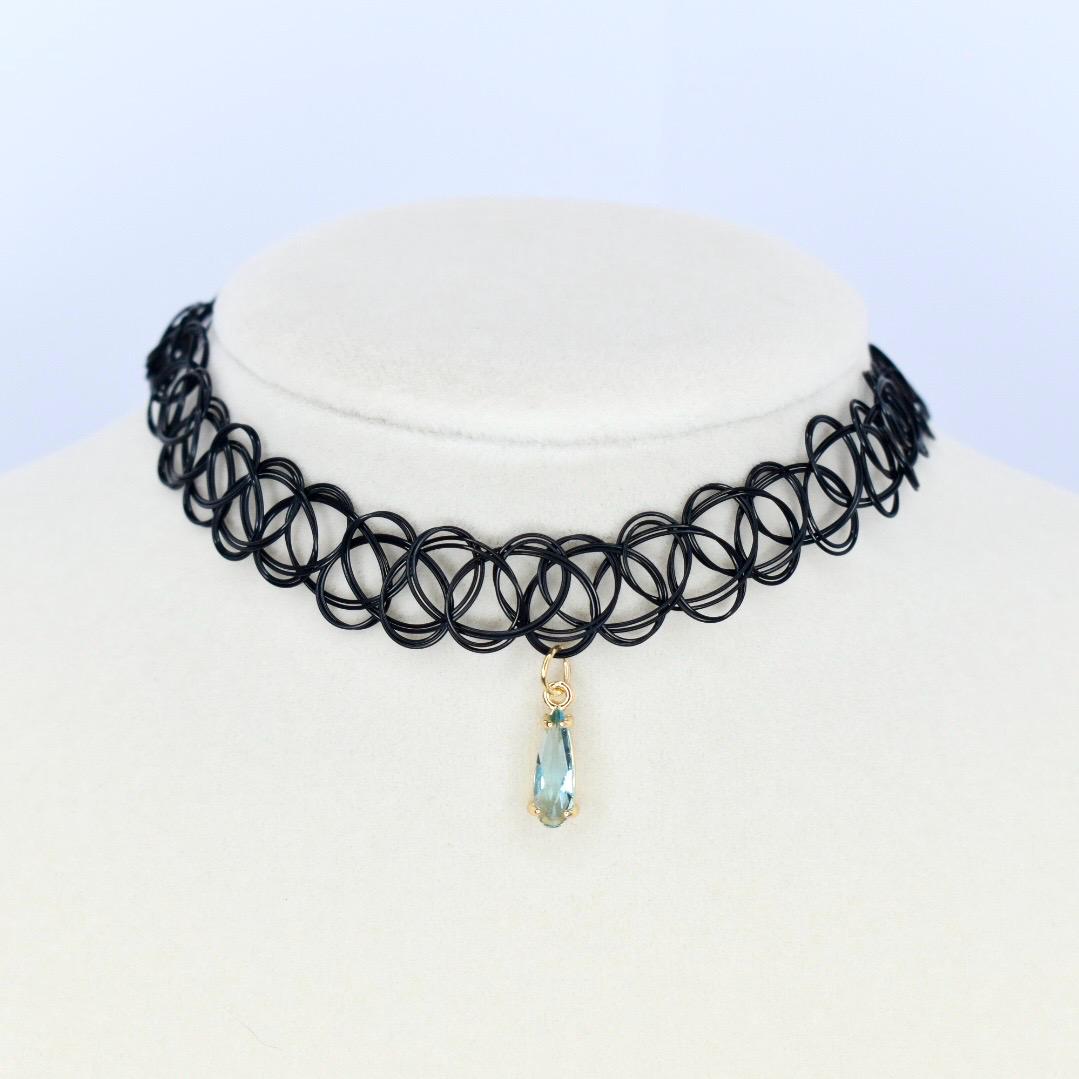 A black choker necklace with a pendant. The necklace is made of small, interconnected circles. A teardrop-shaped pendant hangs from the center, featuring a light blue gemstone set in a gold-colored metal.