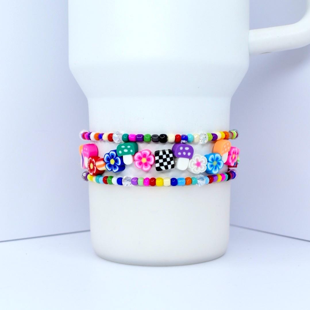 Three colorful beaded bracelets with flower, mushroom, and checkerboard designs are wrapped around a white tumbler. The bracelets feature a variety of bright colors, including pink, blue, green, and yellow.