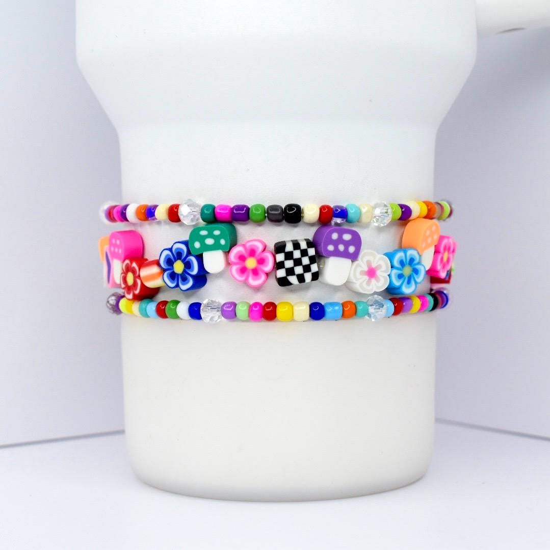 Three colorful beaded bracelets with flower, mushroom, and checkerboard charms. The bracelets feature a variety of bright colors, including pink, blue, green, and orange. The bracelets are stacked on top of each other.