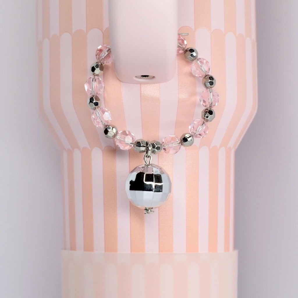 A pink and white striped tumbler with a beaded handle charm bracelet featuring clear pink beads and silver accents. A small, mirrored disco ball charm hangs from the bracelet, adding a touch of sparkle.