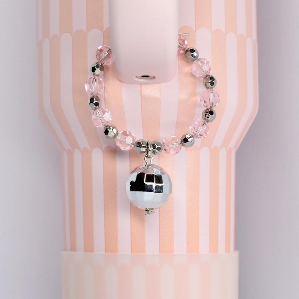 A pink and white striped tumbler with a beaded handle charm bracelet featuring clear pink beads and silver accents. A small, mirrored disco ball charm hangs from the bracelet, adding a touch of sparkle.