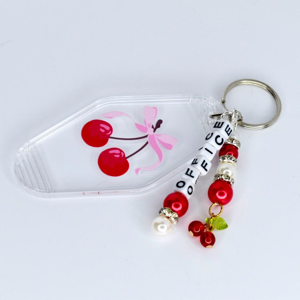 A clear acrylic keychain with a cherry design. The keychain features a transparent base with a cherry graphic and the word 'OFFICE' spelled out in white letter beads. Red beads and faux pearls are strung on the keychain with a metal ring.