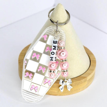 A clear acrylic keychain with a checkered pattern of white squares and pink bows. The keychain has the word 'HOME' spelled out in white letter beads. Pink pearl beads and silver accents are attached, along with a silver bow charm. The keychain is on a wooden stand.