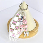 A clear acrylic keychain with a checkered pattern of white squares and pink bows. The keychain has the word 'HOME' spelled out in white letter beads. Pink pearl beads and silver accents are attached, along with a silver bow charm. The keychain is on a wooden stand.