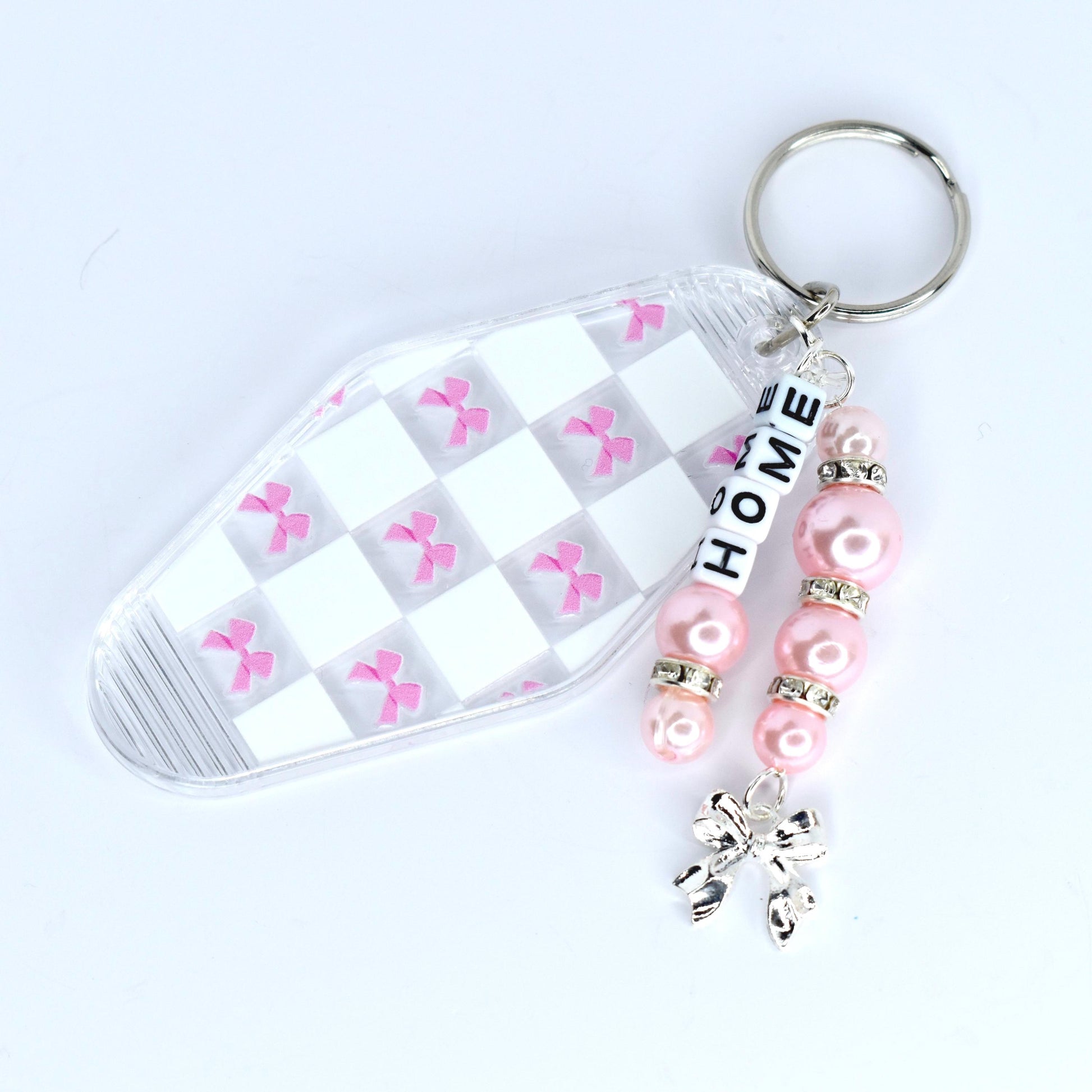 A clear motel-style key tag with a white and pink checkered pattern and pink bow accents. The tag is attached to a silver keyring with a beaded charm. The charm features pink beads, silver accents, and the word 'HOME' spelled out in white letter beads. A silver bow charm dangles at the bottom.