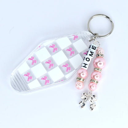 A clear motel-style key tag with a white and pink checkered pattern and pink bow accents. The tag is attached to a silver keyring with a beaded charm. The charm features pink beads, silver accents, and the word 'HOME' spelled out in white letter beads. A silver bow charm dangles at the bottom.