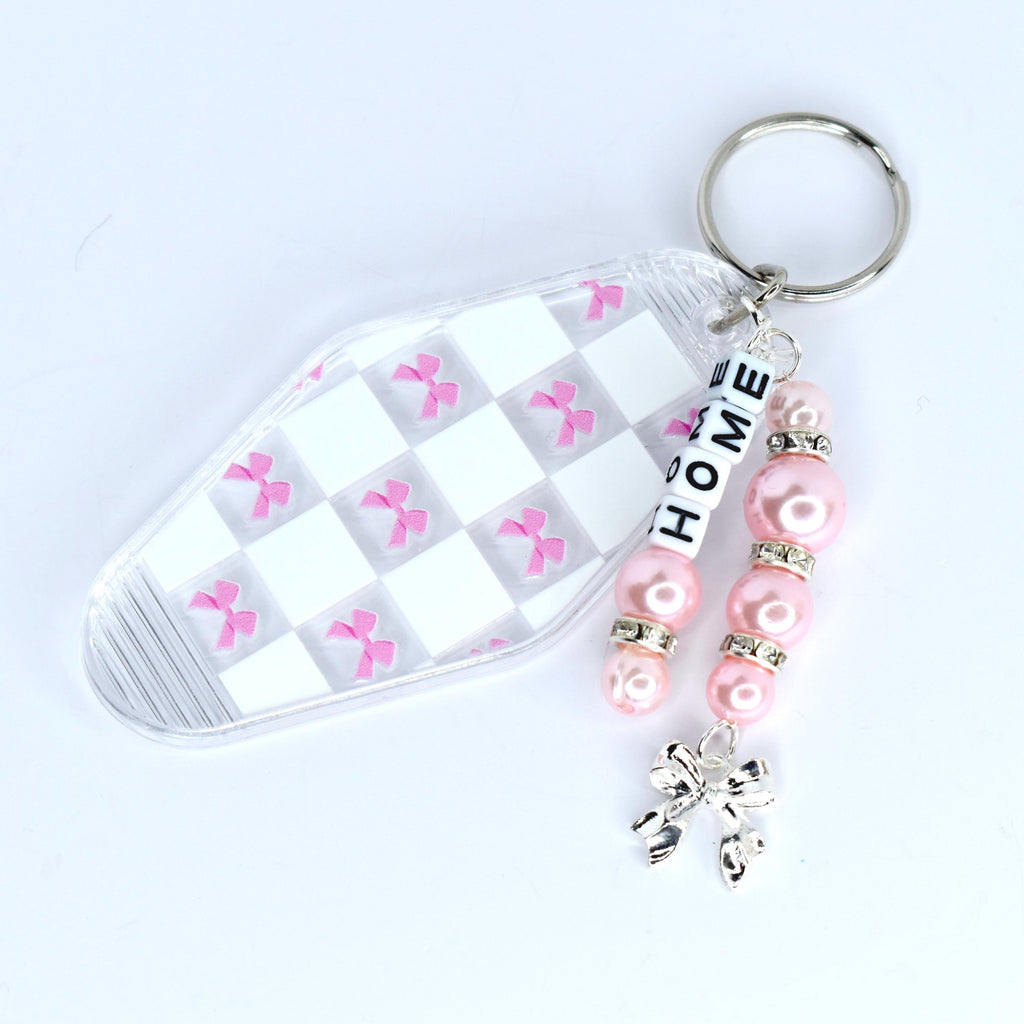 A clear motel-style key tag with a white and pink checkered pattern and pink bow accents. The tag is attached to a silver keyring with a beaded charm. The charm features pink beads, silver accents, and the word 'HOME' spelled out in white letter beads. A silver bow charm dangles at the bottom.