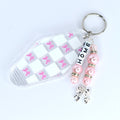 A clear motel-style key tag with a white and pink checkered pattern and pink bow accents. The tag is attached to a silver keyring with a beaded charm. The charm features pink beads, silver accents, and the word 'HOME' spelled out in white letter beads. A silver bow charm dangles at the bottom.