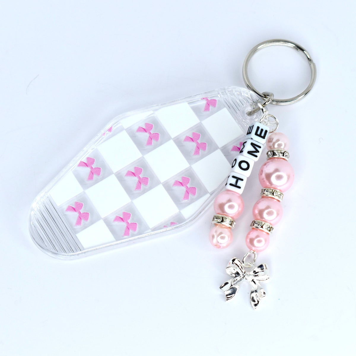 A clear motel-style key tag with a white and pink checkered pattern and pink bow accents. The tag is attached to a silver keyring with a beaded charm. The charm features pink beads, silver accents, and the word 'HOME' spelled out in white letter beads. A silver bow charm dangles at the bottom.