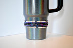 A silver glitter tumbler with a black handle. A decorative band wraps around the middle of the tumbler, featuring rows of white and blue beads, with a center row of black beads and black arrow-shaped accents. The tumbler sits on a white surface.