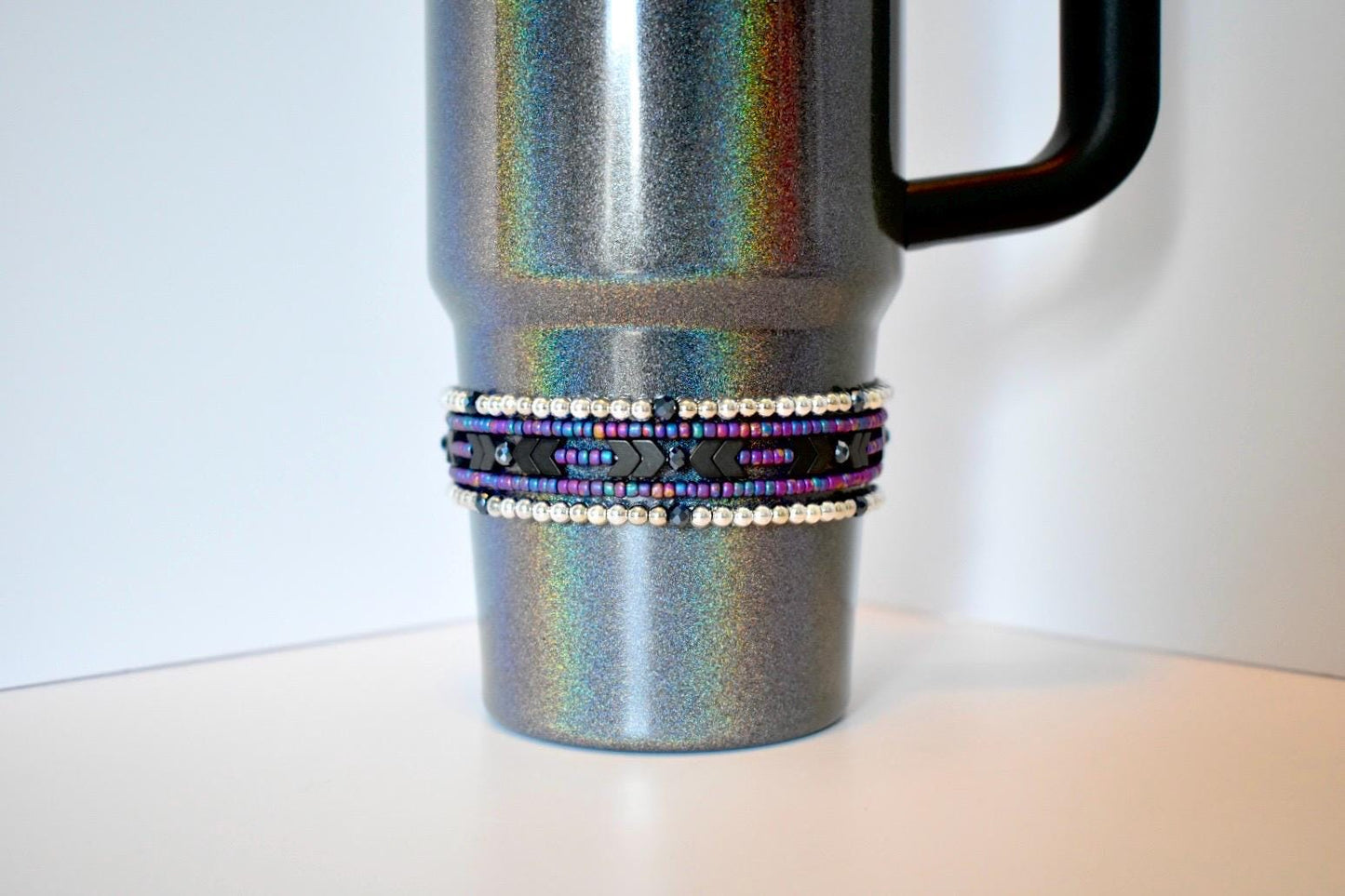 A silver glitter tumbler with a black handle. A decorative band wraps around the middle of the tumbler, featuring rows of white and blue beads, with a center row of black beads and black arrow-shaped accents. The tumbler sits on a white surface.