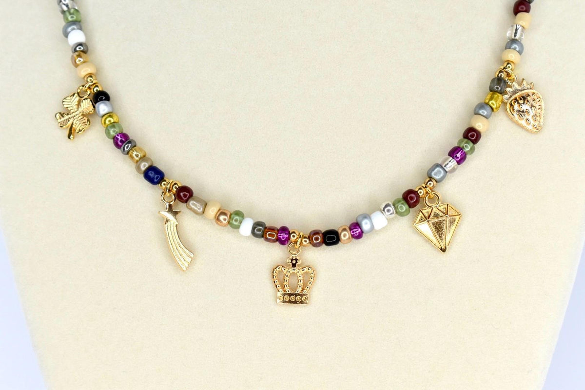 A beaded necklace with a variety of colorful beads and gold-colored charms. The charms include a crown, a diamond, a shooting star, a strawberry, and a four-leaf clover. The necklace is displayed on a white surface.