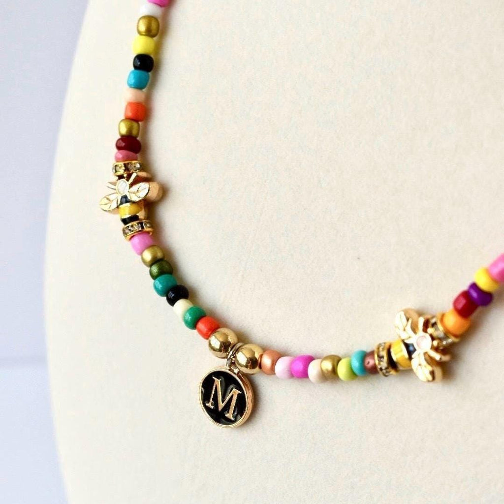 A beaded necklace featuring a sequence of multicolored small beads, two gold-toned bee charms with clear accents, and a circular black pendant with a gold-colored letter M centered on the front, suspended from a gold-toned jump ring.