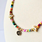 A beaded necklace featuring a sequence of multicolored small beads, two gold-toned bee charms with clear accents, and a circular black pendant with a gold-colored letter M centered on the front, suspended from a gold-toned jump ring.