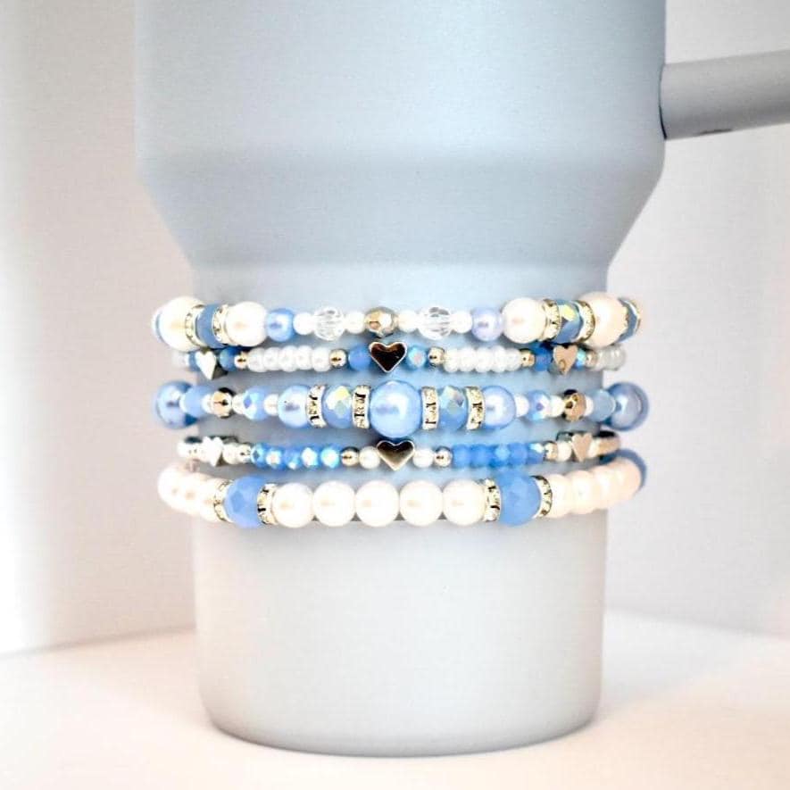 A set of five beaded tumbler bracelets wrapped around a matte light blue Stanley-style tumbler. The bracelets feature shades of blue, pearl white, and clear beads with silver and gold accents, including small heart and rhinestone spacer beads, creating a serene and dreamy aesthetic.