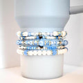 A set of five beaded tumbler bracelets wrapped around a matte light blue Stanley-style tumbler. The bracelets feature shades of blue, pearl white, and clear beads with silver and gold accents, including small heart and rhinestone spacer beads, creating a serene and dreamy aesthetic.