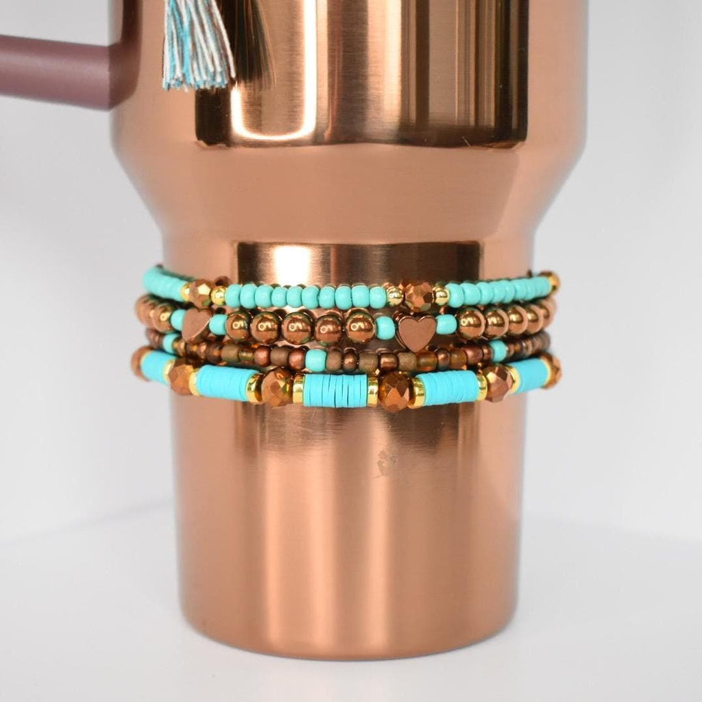 A stack of beaded bracelets in turquoise, gold, and brown tones. The bracelets are wrapped around a copper-colored tumbler. Some bracelets feature heart-shaped beads. The tumbler has a tassel attached to the handle.