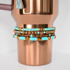 A stack of beaded bracelets in turquoise, gold, and brown tones. The bracelets are wrapped around a copper-colored tumbler. Some bracelets feature heart-shaped beads. The tumbler has a tassel attached to the handle.
