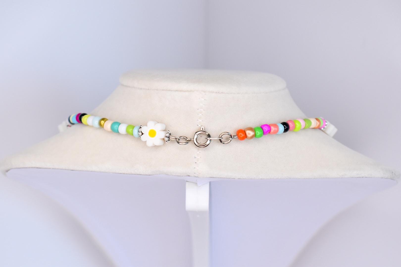 A colorful beaded necklace with a daisy pendant. The necklace features a variety of small, round beads in shades of white, yellow, green, blue, pink, orange, and black. The daisy pendant is white with a yellow center. The necklace is fastened with a silver clasp.