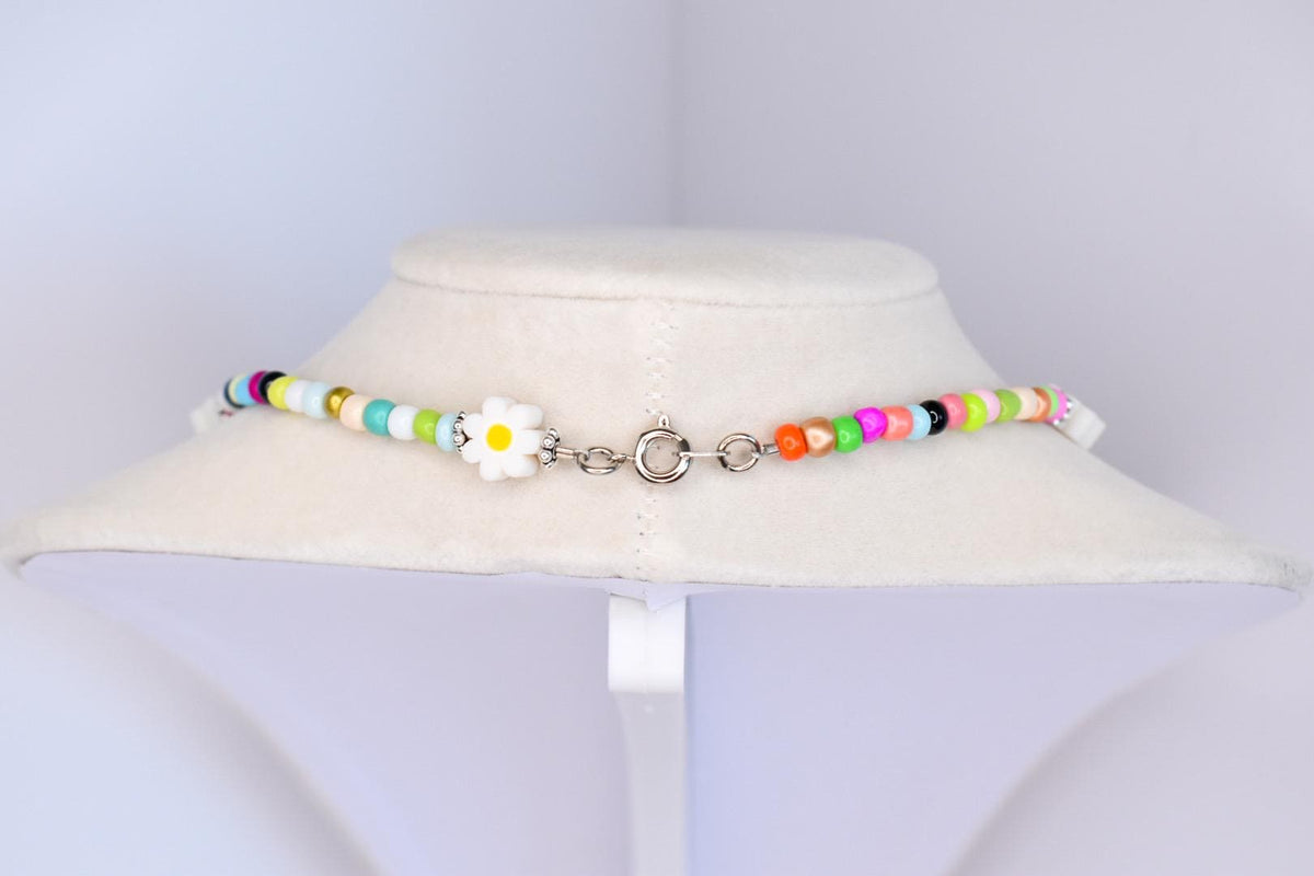 A colorful beaded necklace with a daisy pendant. The necklace features a variety of small, round beads in shades of white, yellow, green, blue, pink, orange, and black. The daisy pendant is white with a yellow center. The necklace is fastened with a silver clasp.