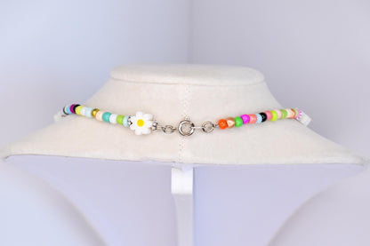 A colorful beaded necklace with a daisy pendant. The necklace features a variety of small, round beads in shades of white, yellow, green, blue, pink, orange, and black. The daisy pendant is white with a yellow center. The necklace is fastened with a silver clasp.