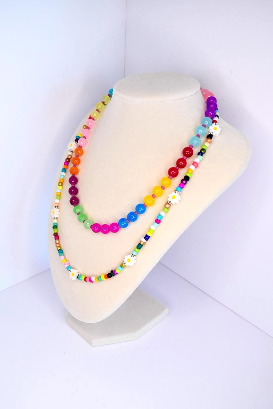 Two colorful beaded necklaces displayed on a white mannequin. The necklaces feature a variety of bright beads including red, yellow, green, blue, and pink. One necklace has daisy-shaped beads. The necklaces are a vibrant accessory, perfect for adding a pop of color.