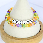 Two beaded bracelets with the word "EVOLVE" spelled out in white letter beads. The bracelets feature colorful beads in shades of orange, yellow, green, purple, and blue, with gold accents. The bracelets are displayed on a white cone-shaped stand.